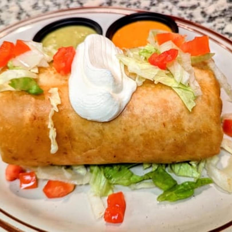 A golden-brown chimichanga topped with sour cream, lettuce, and diced tomatoes, with two dipping sauces.